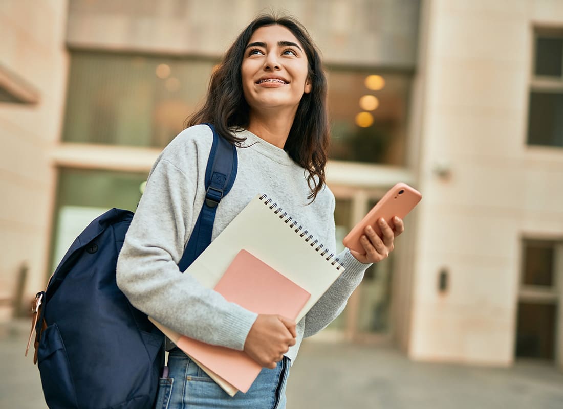Student Health Insurance - Portrait of a Young Girl Holding Books and Wearing a Backpack Standing in Front of a School Building While Holding her Phone